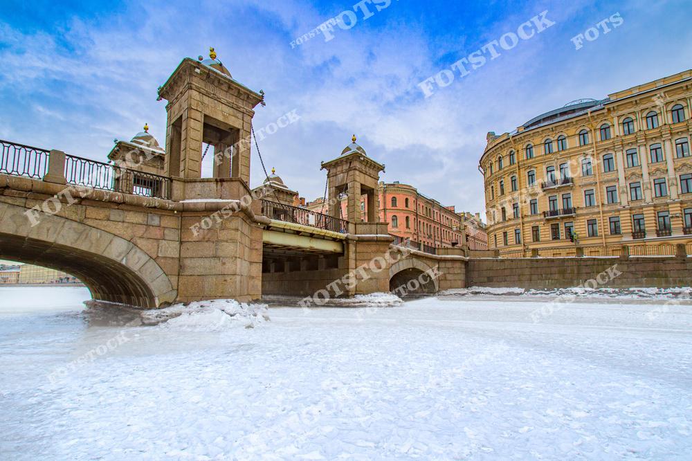 На фотографии изображен мост Ломоносова, один из красивейших мостов Санкт-Петербурга, в зимний день. Мост, выполненный в неоклассическом стиле, выглядит особенно величественно на фоне заснеженной реки и голубого неба. Зимняя сцена с ледяным покровом подчеркивает историческое наследие и красоту архитектурных элементов моста, а также гармонично сочетается с городской атмосферой.